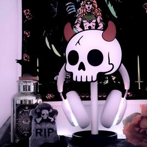 May include: A white skull-shaped headphone stand with red horns, holding white headphones with glowing earcups. A decorative tombstone with "RIP" is in front of a black background with a gothic illustration.