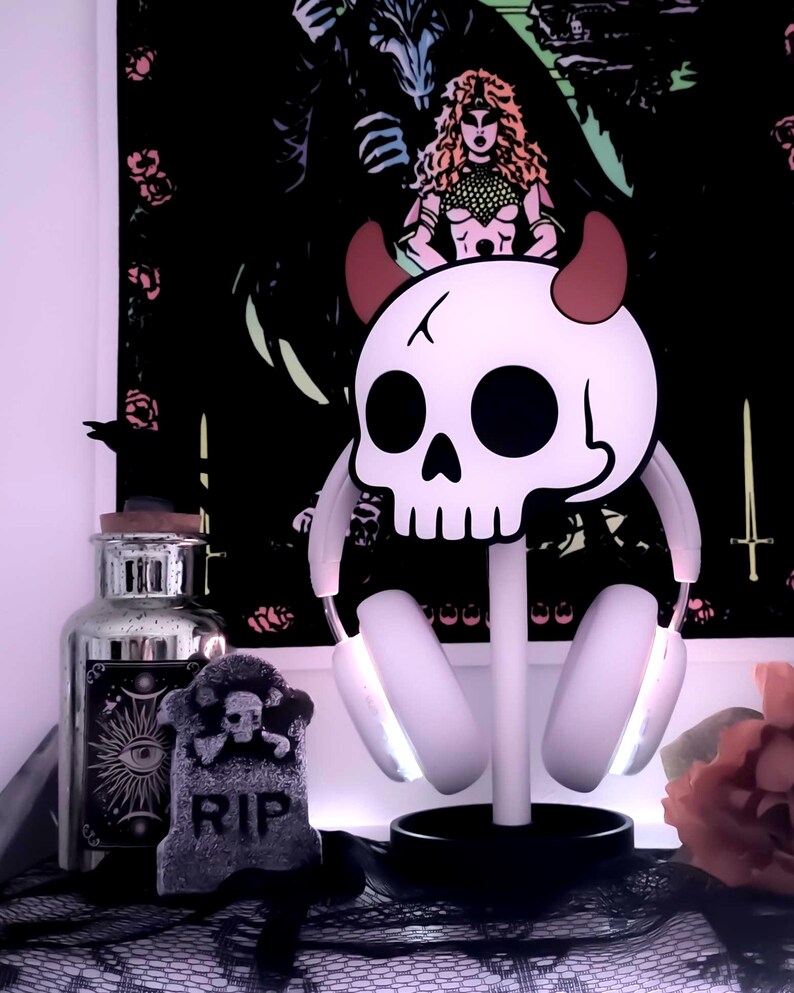 May include: A white skull-shaped headphone stand with red horns, holding white headphones with glowing earcups. A decorative tombstone with "RIP" is in front of a black background with a gothic illustration.