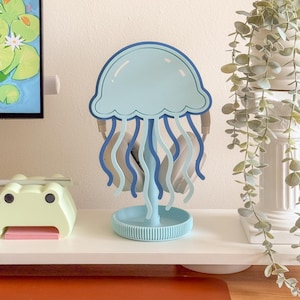 May include: A light blue and dark blue jellyfish-shaped headphone stand. The stand has a round base and a top shaped like a jellyfish. Headphones are hanging from the stand. The stand is on a white surface.
