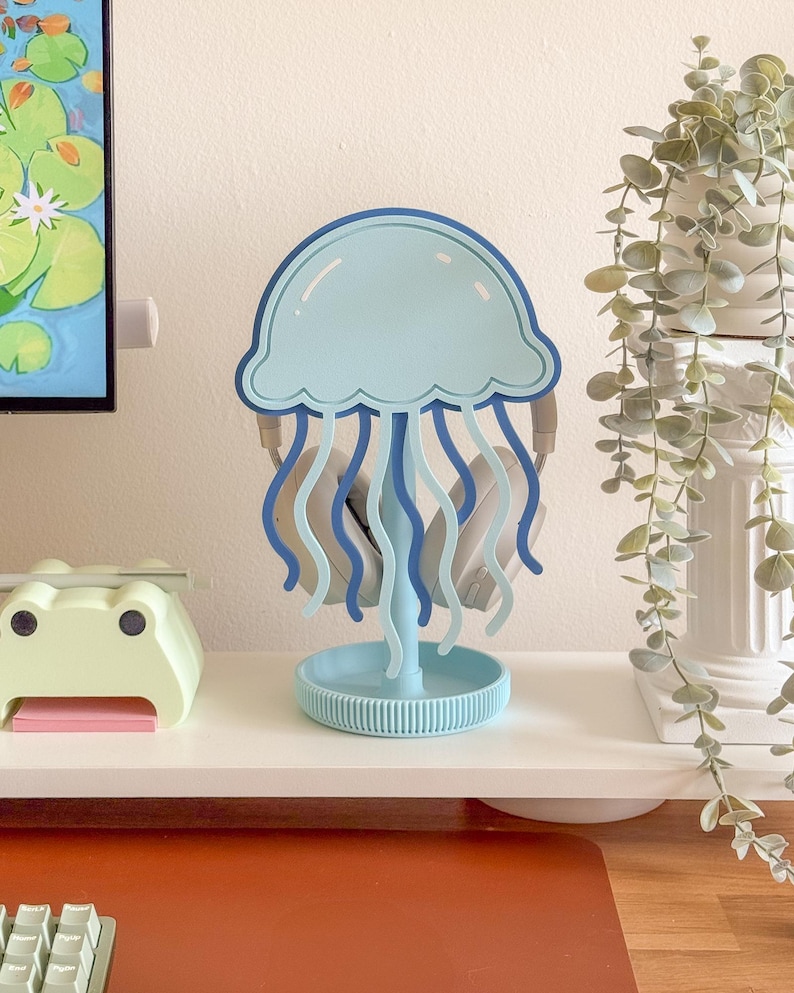 May include: A light blue and dark blue jellyfish-shaped headphone stand. The stand has a round base and a top shaped like a jellyfish. Headphones are hanging from the stand. The stand is on a white surface.