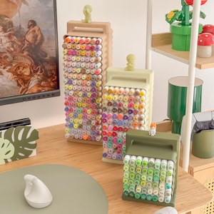 May include: Three decorative marker holders in beige, light green, and olive green, each filled with colorful markers. The holders have a handle and a textured design. Small figurines sit atop each holder.