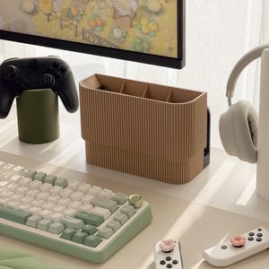 May include: A desk setup with a brown ribbed organiser, a white and green keyboard, and a black game controller. A pair of white headphones and two white game controllers with pink flower accents are also visible.