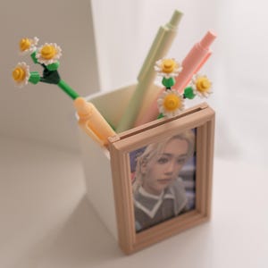Pu&ograve; includere: Un contenitore quadrato bianco con penne e una piccola foto incorniciata. Le penne sono di colori pastello e hanno accenti di fiori di margherita. La foto &egrave; di una persona con i capelli biondi. La cornice &egrave; marrone chiaro.