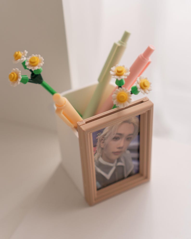 Pu&ograve; includere: Un contenitore quadrato bianco con penne e una piccola foto incorniciata. Le penne sono di colori pastello e hanno accenti di fiori di margherita. La foto &egrave; di una persona con i capelli biondi. La cornice &egrave; marrone chiaro.