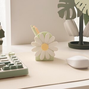 Pu&ograve; includere: Un portapenne a forma di margherita verde chiaro con petali bianchi e un centro giallo. L'accessorio da scrivania contiene penne e si trova su una scrivania bianca con tastiera e mouse abbinati. Un supporto decorativo per cuffie a forma di foglia di monstera &egrave; sullo sfondo.