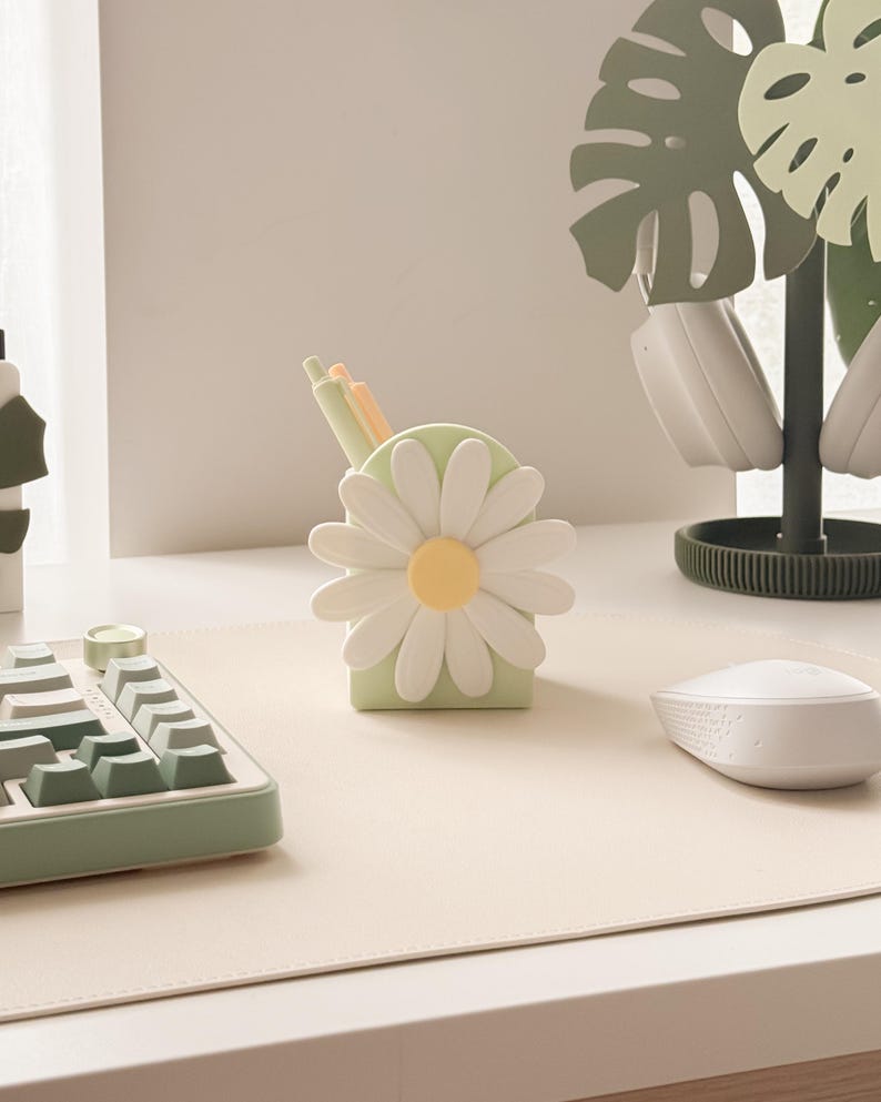 Pu&ograve; includere: Un portapenne a forma di margherita verde chiaro con petali bianchi e un centro giallo. L'accessorio da scrivania contiene penne e si trova su una scrivania bianca con tastiera e mouse abbinati. Un supporto decorativo per cuffie a forma di foglia di monstera &egrave; sullo sfondo.