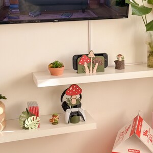 Pu&ograve; includere: Esposizione di oggetti decorativi su mensole bianche. Sono visibili un supporto per console Switch con disegni di funghi, una piccola succulenta in un vaso di terracotta e una statuina di fungo in legno. &Egrave; presente anche un supporto per cuffie da gioco con design a fungo e un controller bianco.