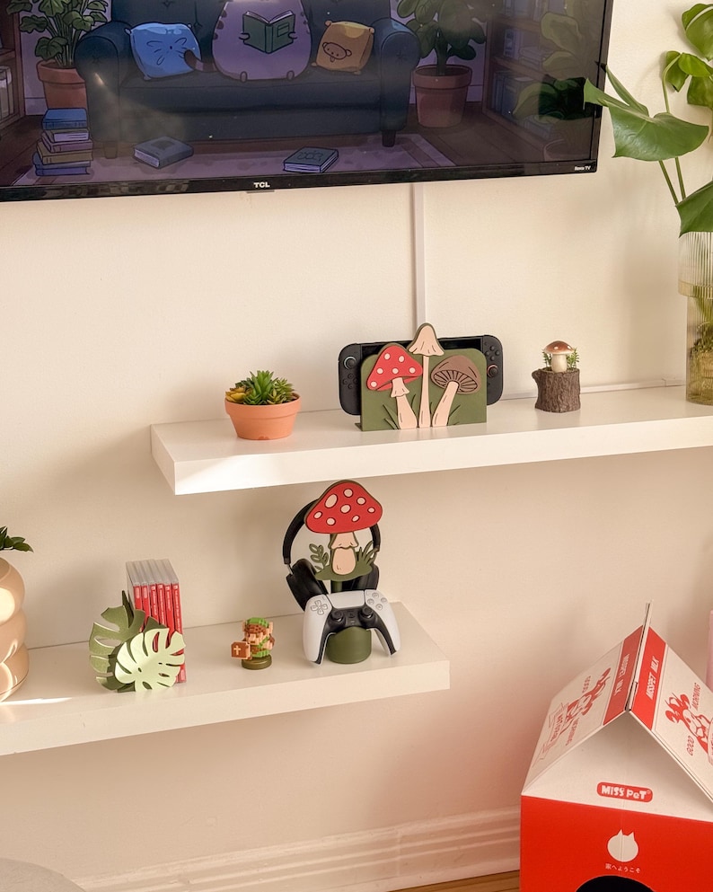 Pu&ograve; includere: Esposizione di oggetti decorativi su mensole bianche. Sono visibili un supporto per console Switch con disegni di funghi, una piccola succulenta in un vaso di terracotta e una statuina di fungo in legno. &Egrave; presente anche un supporto per cuffie da gioco con design a fungo e un controller bianco.