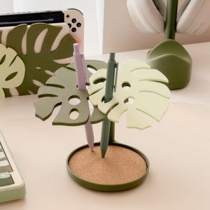 Pu&ograve; includere: Un portapenne decorativo a forma di foglia di monstera verde con base in sughero. Contiene una penna lilla e una verde. Altri oggetti nell'immagine includono una custodia per console Switch e cuffie.