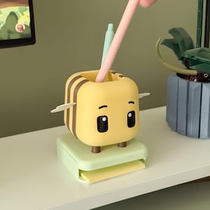 May include: A yellow and brown bee-shaped desk organizer with a light green base and a small drawer. The organizer has two black eyes and two small wings. A pink pen and a light green pen are inside the organizer.