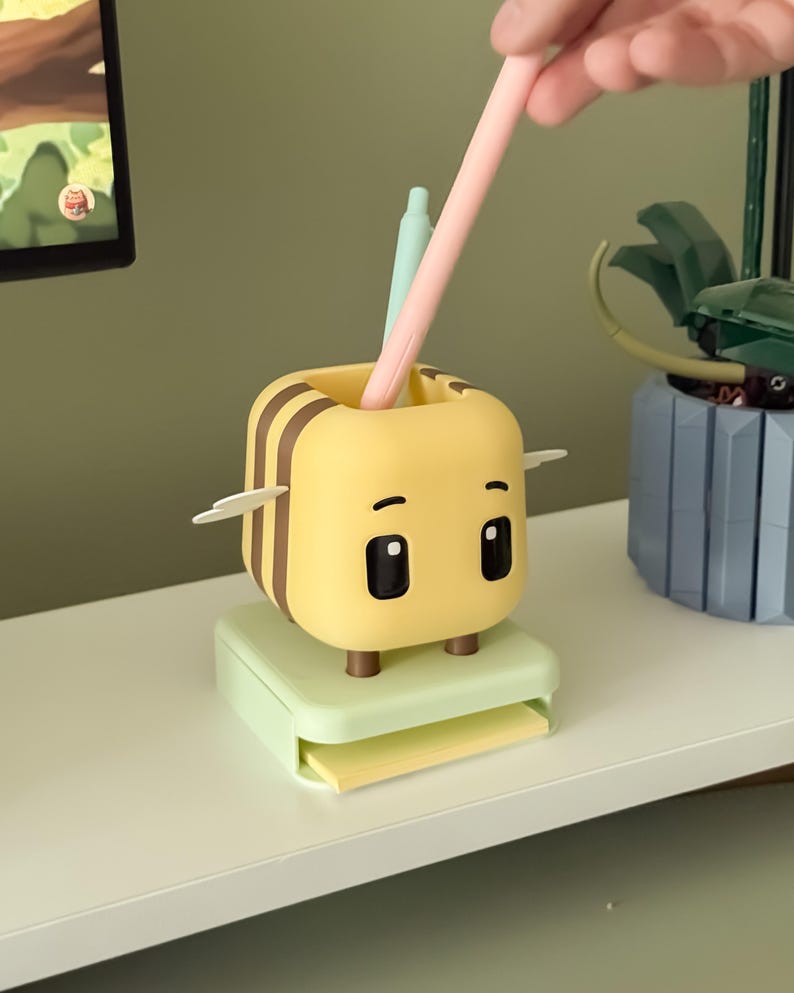 May include: A yellow and brown bee-shaped desk organizer with a light green base and a small drawer. The organizer has two black eyes and two small wings. A pink pen and a light green pen are inside the organizer.