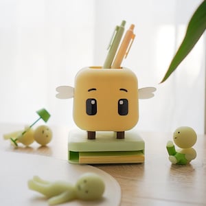 May include: A whimsical yellow pen holder with a cartoon face, small wings, and black eyes. It sits on a light green base with a stack of yellow sticky notes. Two pens are in the holder, and small figurines are scattered around.