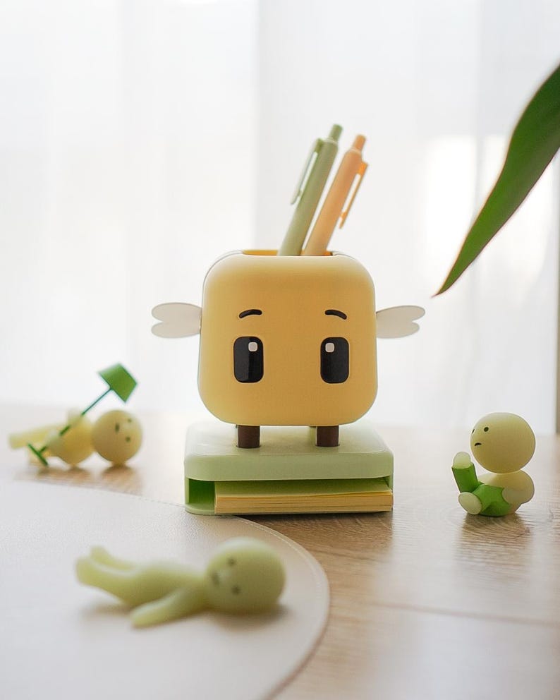 May include: A whimsical yellow pen holder with a cartoon face, small wings, and black eyes. It sits on a light green base with a stack of yellow sticky notes. Two pens are in the holder, and small figurines are scattered around.