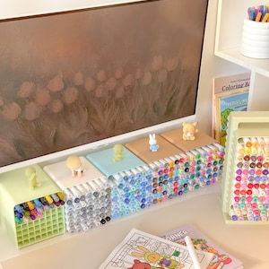 May include: A collection of square marker organizers in pastel colors, each topped with a small character figurine. The organizers hold numerous colored markers. A coloring book with a cartoon illustration and a marker stand are also visible.