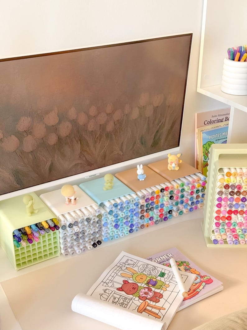 May include: A collection of square marker organizers in pastel colors, each topped with a small character figurine. The organizers hold numerous colored markers. A coloring book with a cartoon illustration and a marker stand are also visible.