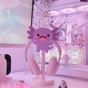 May include: A white headset rests on a decorative stand featuring a purple axolotl design. The stand is on a white surface, with a small penguin figurine and other decorative items in the background. The overall aesthetic is a soft, pastel pink.