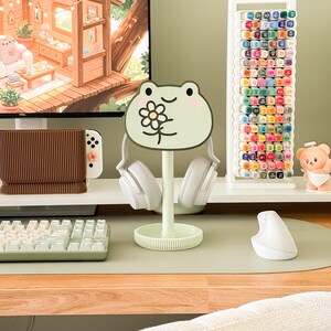 May include: A light green frog-shaped headphone stand with a flower design, holding white headphones. The stand sits on a desk with a keyboard, mouse, and a white marker holder filled with colorful markers. A computer monitor displays a cartoon scene.