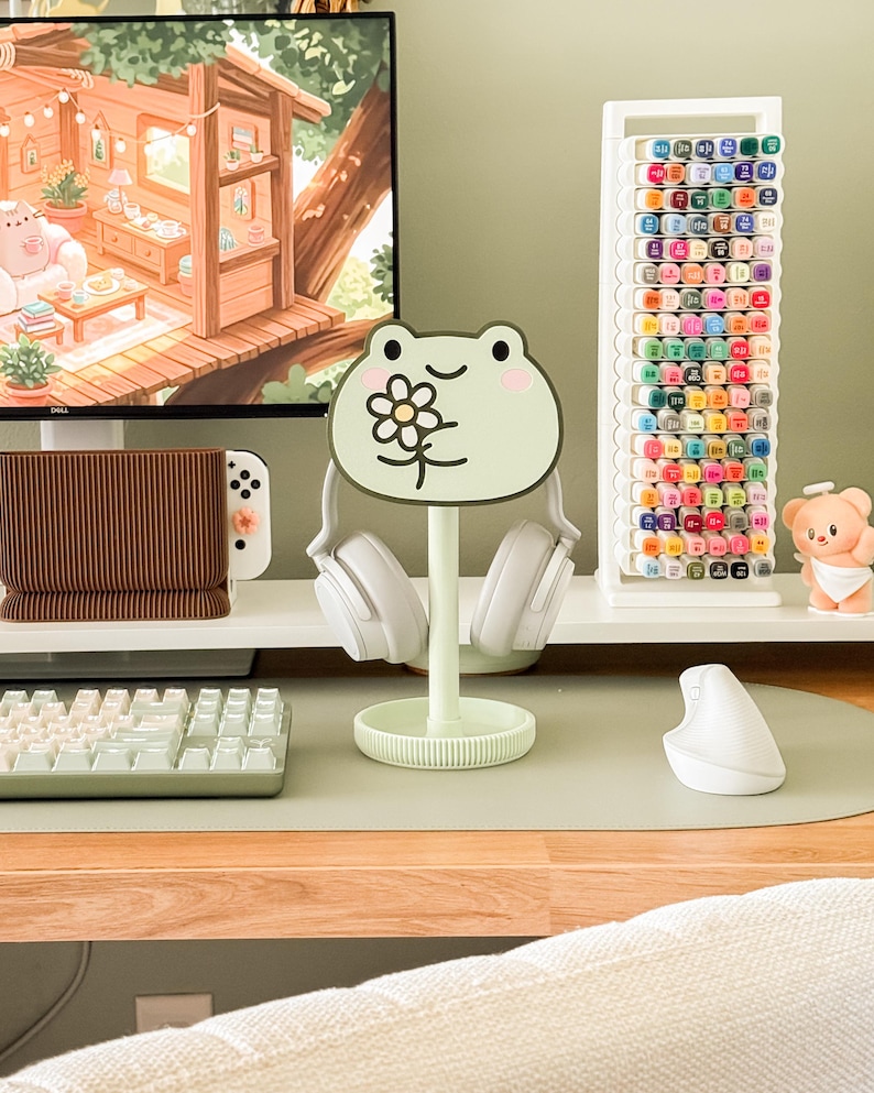 May include: A light green frog-shaped headphone stand with a flower design, holding white headphones. The stand sits on a desk with a keyboard, mouse, and a white marker holder filled with colorful markers. A computer monitor displays a cartoon scene.