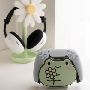 May include: A white and black headset rests on a daisy-shaped stand with a light green base. A white and gray game controller with a green frog design is in the foreground. The frog has a flower on its chest.