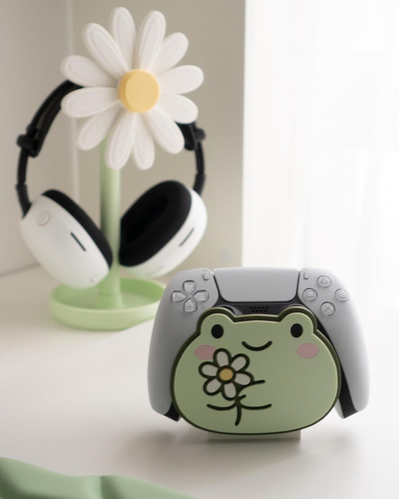 May include: A white and black headset rests on a daisy-shaped stand with a light green base. A white and gray game controller with a green frog design is in the foreground. The frog has a flower on its chest.