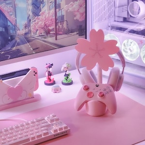 May include: Pink gaming desk setup with a pink cherry blossom-shaped headset stand, a pink Xbox controller, a pink keyboard, and a pink Nintendo Switch dock.