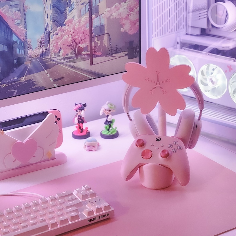 May include: Pink gaming desk setup with a pink cherry blossom-shaped headset stand, a pink Xbox controller, a pink keyboard, and a pink Nintendo Switch dock.