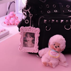 May include: A black handbag with silver studs and grommets, a pink framed photo, and a fluffy pink doll with a bib that says "Colors" sit on a pink surface. A stack of pink Kirby figurines is in the background.