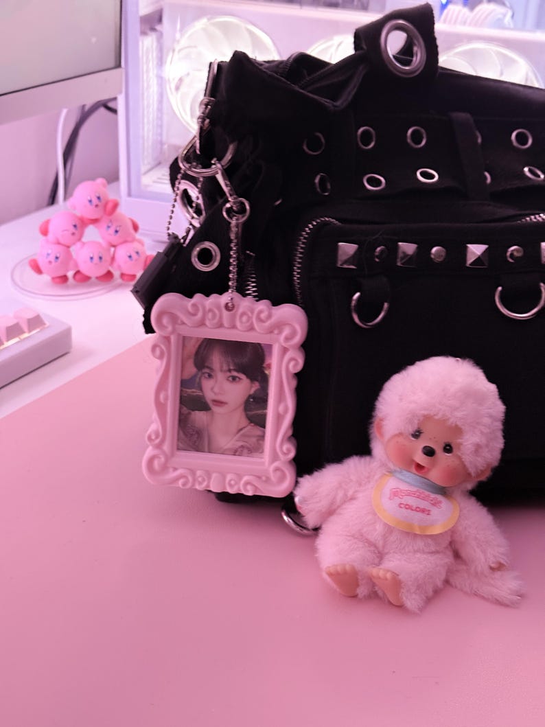 May include: A black handbag with silver studs and grommets, a pink framed photo, and a fluffy pink doll with a bib that says "Colors" sit on a pink surface. A stack of pink Kirby figurines is in the background.