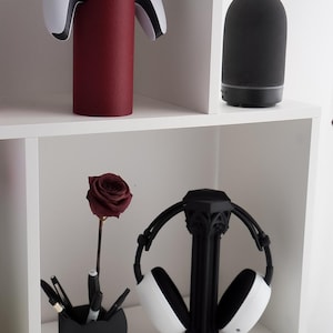 May include: A white and black gaming controller rests on a cylindrical, textured, burgundy stand. A black essential oil diffuser and a black headphone stand with white headphones are also visible. A black pen holder with pens and a burgundy rose complete the display.