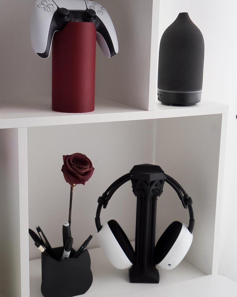 May include: A white and black gaming controller rests on a cylindrical, textured, burgundy stand. A black essential oil diffuser and a black headphone stand with white headphones are also visible. A black pen holder with pens and a burgundy rose complete the display.