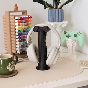 Puede incluir: Una composici&oacute;n de escritorio con un soporte negro para auriculares que sostiene auriculares blancos. Tambi&eacute;n se ven un organizador de marcadores beige, una taza verde y un mando de juegos verde menta. Una maceta azul con una planta descansa sobre un pedestal blanco.