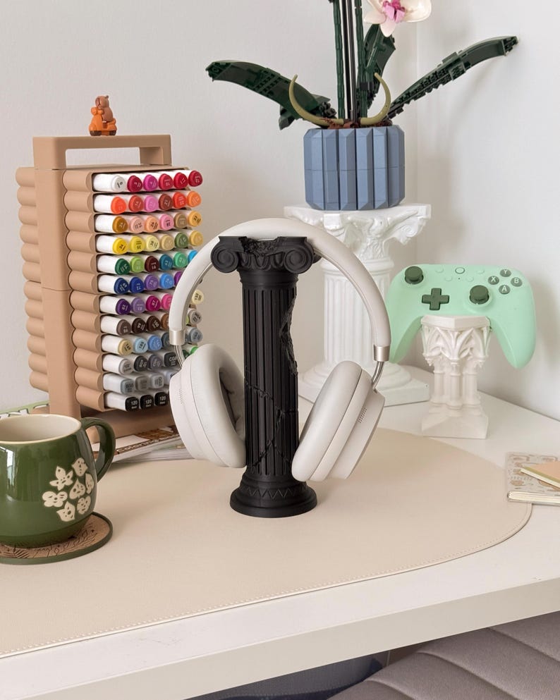Puede incluir: Una composici&oacute;n de escritorio con un soporte negro para auriculares que sostiene auriculares blancos. Tambi&eacute;n se ven un organizador de marcadores beige, una taza verde y un mando de juegos verde menta. Una maceta azul con una planta descansa sobre un pedestal blanco.