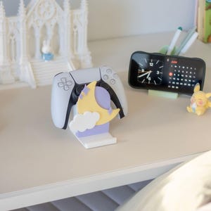 May include: A white gaming controller rests on a stand featuring a light purple rectangular base, a yellow crescent moon, a white cloud, and small purple star accents. The stand is designed to hold the controller upright on a flat surface.
