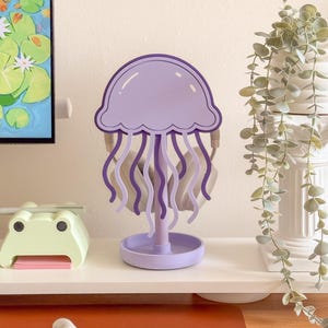 May include: A purple jellyfish-shaped headphone stand. The stand features a round base, a central support, and a jellyfish head with flowing tentacles. Headphones are hanging on the stand. The stand is on a white shelf with a frog-shaped item and a plant.