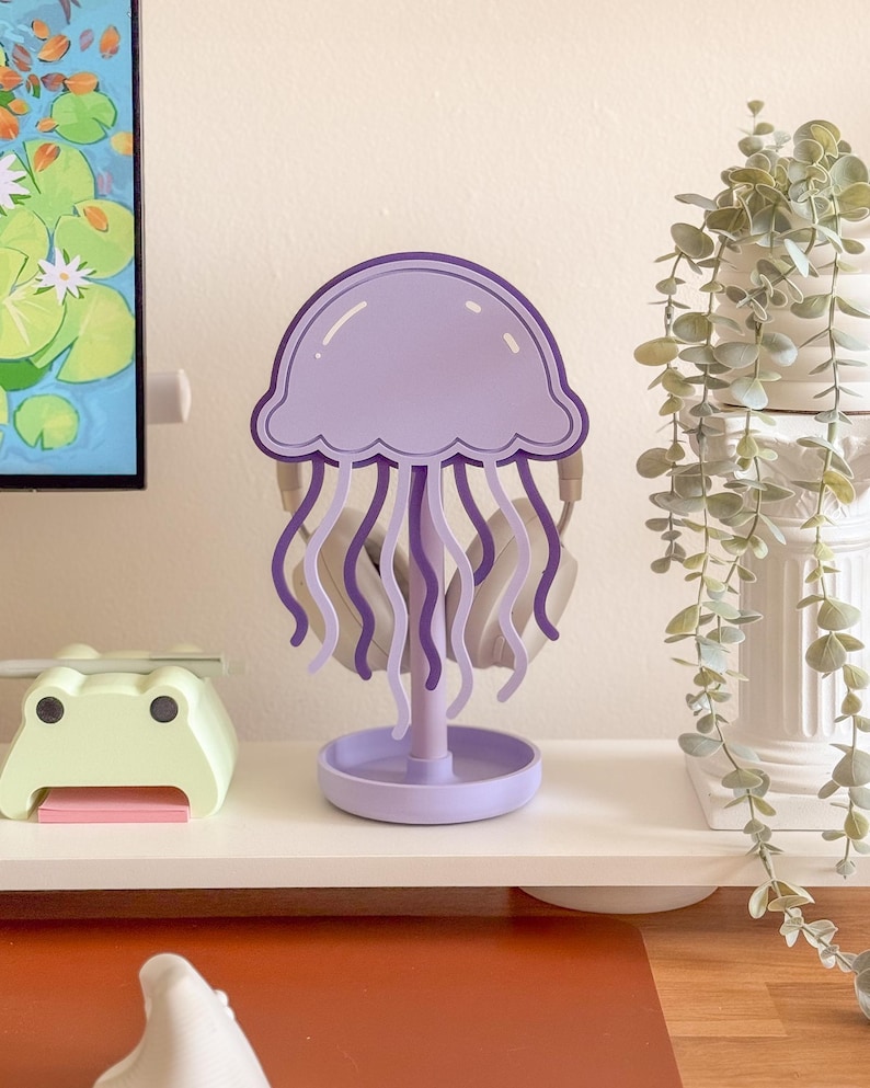 May include: A purple jellyfish-shaped headphone stand. The stand features a round base, a central support, and a jellyfish head with flowing tentacles. Headphones are hanging on the stand. The stand is on a white shelf with a frog-shaped item and a plant.