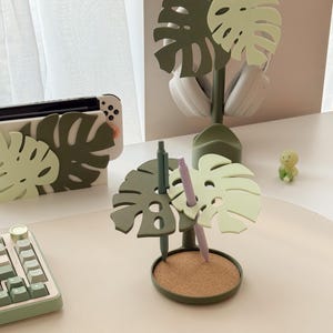 Pu&ograve; includere: Un organizer da scrivania a forma di foglia di monstera verde con base in sughero, per penne. Sono visibili anche un supporto per cuffie e una custodia per Switch abbinati. La tavolozza dei colori include verdi e un beige chiaro.