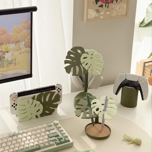 Pu&ograve; includere: Una scrivania con una Nintendo Switch, una tastiera e un controller PlayStation. Gli accessori includono un portapenne a forma di foglia di monstera, un supporto per Switch e un supporto per controller, tutti in tonalit&agrave; verdi.