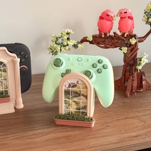 May include: A light green wireless video game controller resting on a peach-coloured stand shaped like an arched window with a garden scene print. The stand features a small planter box base with green texture, positioned near a brick-built tree.
