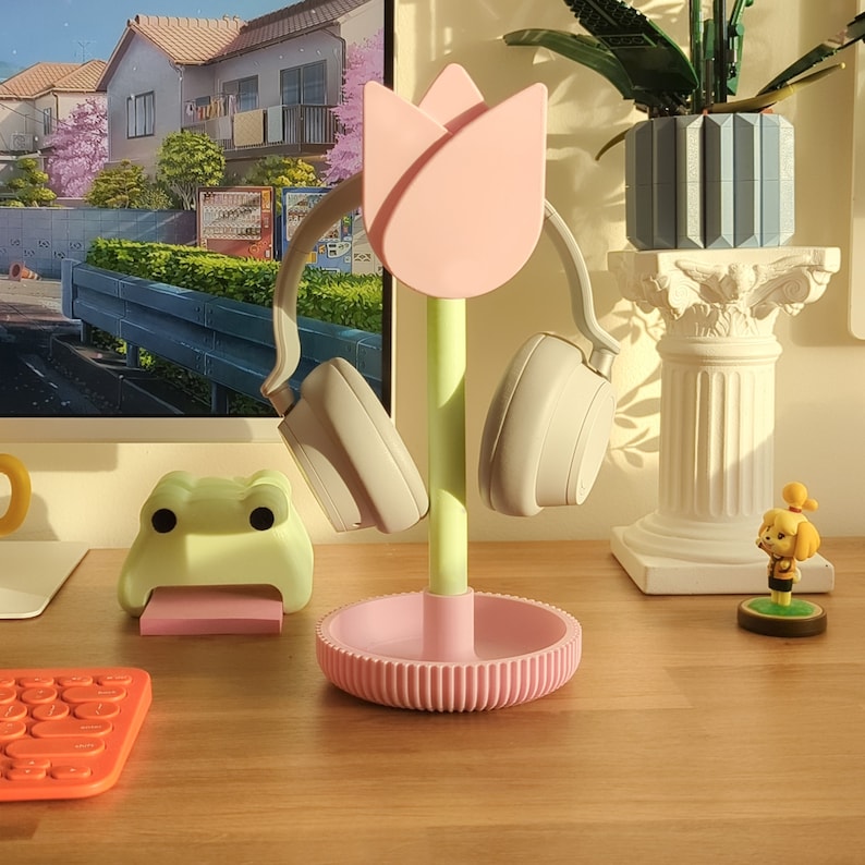 Puede incluir: Un soporte para auriculares en forma de tulip&aacute;n rosa con un tallo verde. El soporte sostiene unos auriculares negros de diadema. El soporte est&aacute; sobre una mesa de madera.