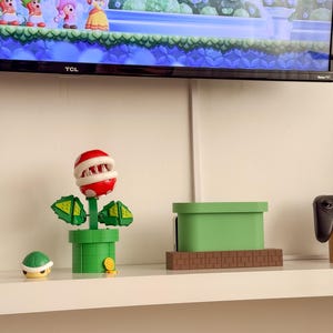 May include: A green and red Piranha Plant toy from the Super Mario Bros. video game series. The toy is made of plastic and has a movable mouth. The toy is on a shelf with a green and brown brick block toy and a green pipe toy.