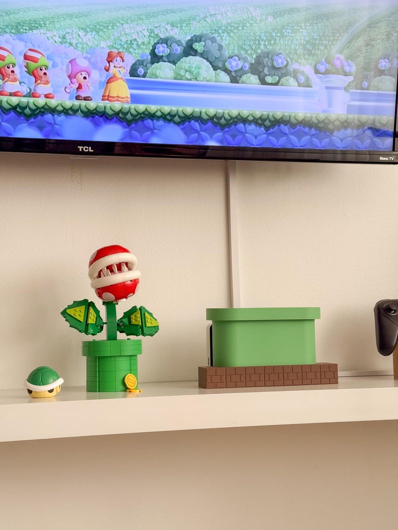 May include: A green and red Piranha Plant toy from the Super Mario Bros. video game series. The toy is made of plastic and has a movable mouth. The toy is on a shelf with a green and brown brick block toy and a green pipe toy.
