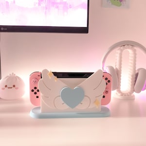 May include: A pink and white Nintendo Switch console stand with a blue heart and white wings. The stand is on a white surface.