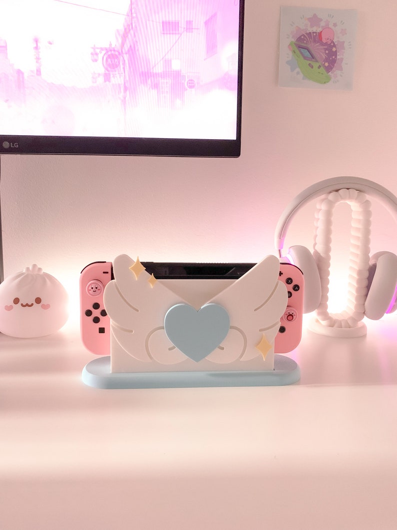 May include: A pink and white Nintendo Switch console stand with a blue heart and white wings. The stand is on a white surface.