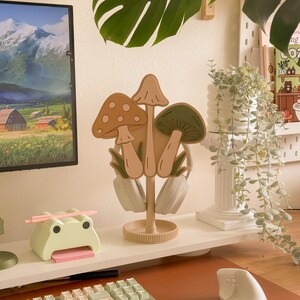 Pu&ograve; includere: Un supporto per cuffie a forma di fungo beige con tre disegni di funghi nei toni del marrone e del verde. Il supporto &egrave; su una mensola bianca con un porta-post-it a forma di rana e una tastiera per computer.