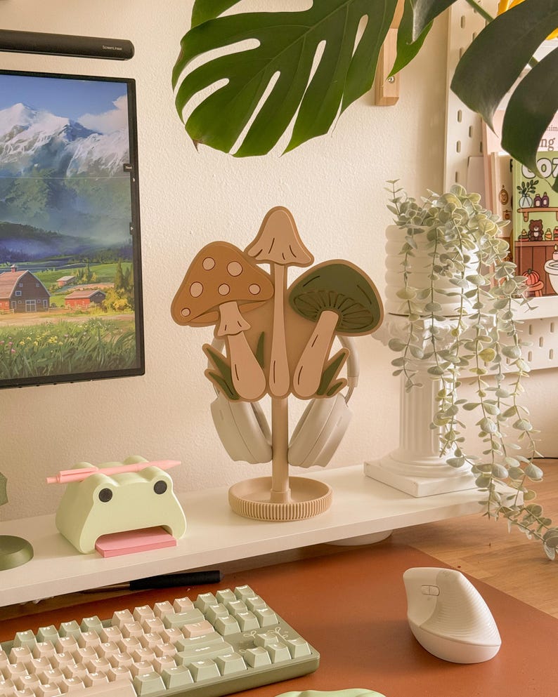 Pu&ograve; includere: Un supporto per cuffie a forma di fungo beige con tre disegni di funghi nei toni del marrone e del verde. Il supporto &egrave; su una mensola bianca con un porta-post-it a forma di rana e una tastiera per computer.