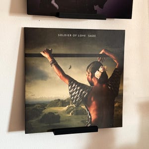 May include: Album cover for Sade's "Soldier of Love" displayed on a wall. The album art features a woman with her back to the viewer, arms raised, with a landscape background. The album title is printed at the top.