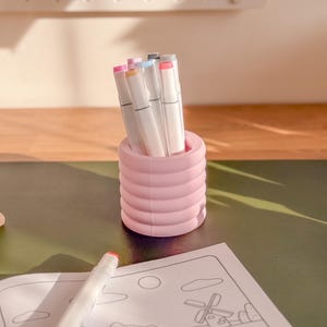 May include: A pink, ribbed pen holder filled with colorful markers. A coloring book with a cartoon illustration is on the table. The markers have white barrels and various colored tips. The pen holder is cylindrical and has a ridged design.