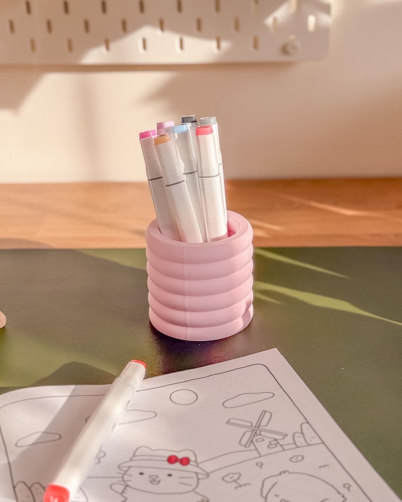 May include: A pink, ribbed pen holder filled with colorful markers. A coloring book with a cartoon illustration is on the table. The markers have white barrels and various colored tips. The pen holder is cylindrical and has a ridged design.