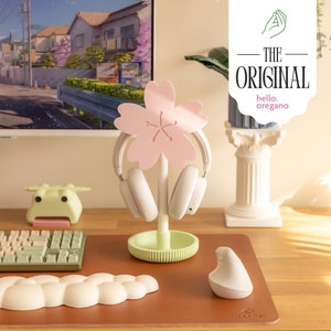 Op de afbeelding: Een roze kersenbloesemvormige koptelefoonstandaard met een groene basis. De standaard houdt een paar witte koptelefoons vast. De standaard staat op een bruin bureau met een wit toetsenbord, een groen kikkerbeeldje en een witte muis.