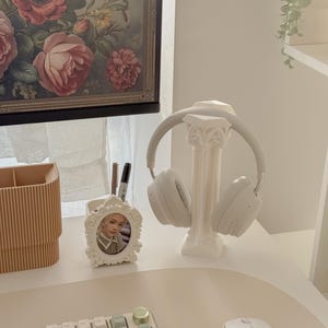 May include: A white desk setup featuring a decorative headphone stand, a framed photo, and a keyboard with mint green keys. A pair of white headphones rests on the stand. A floral painting hangs in the background.
